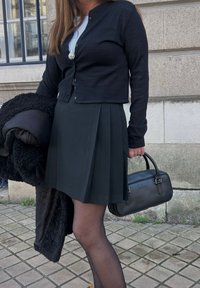 Črn kardigan čez belo bluzo, v kombinaciji s črno naborkano krilo in črnimi nogavicami. Drži črno torbico, z naboranim temnim plaščem, ki ga ima prek roke.