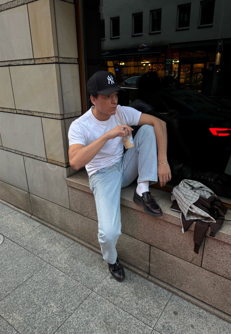 Maglietta bianca, jeans azzurri chiari, cappellino nero e scarpe scure. Seduto su un bordo di pietra con una giacca marrone e una bibita ghiacciata in mano.