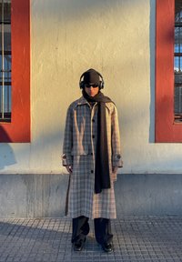 Hosszú bézs és barna kockás kabát nagy gombokkal, fekete sállal és sapkával, fekete nadrággal és fejhallgatóval párosítva.