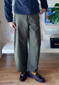 Person wearing olive green wide-leg trousers, dark brown loafers, yellow socks, and a navy jacket standing indoors on a wooden floor.