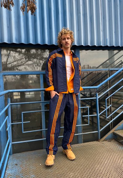 Un conjunto deportivo en color navy y naranja que incluye una chaqueta con rayas naranjas, pantalones a juego y zapatos beige, todo esto frente a un fondo azul con corrugado.