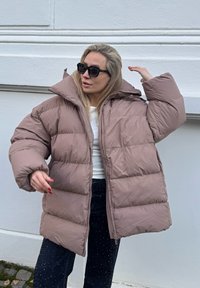 Vatteret jakke i blød tan med høj krave og stor hætte. Har quiltede sektioner og lynlås lukning. B�året over en hvid skjorte og mørke jeans.