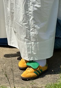 Κίτρινα αθλητικά παπούτσια με πράσινες ρίγες και λεπτομέρειες, υφασμένο σουέτ άνω τμήμα και ιμάντας βελκρό, φορεμένα με λευκές κάλτσες και λευκό ρούχο.