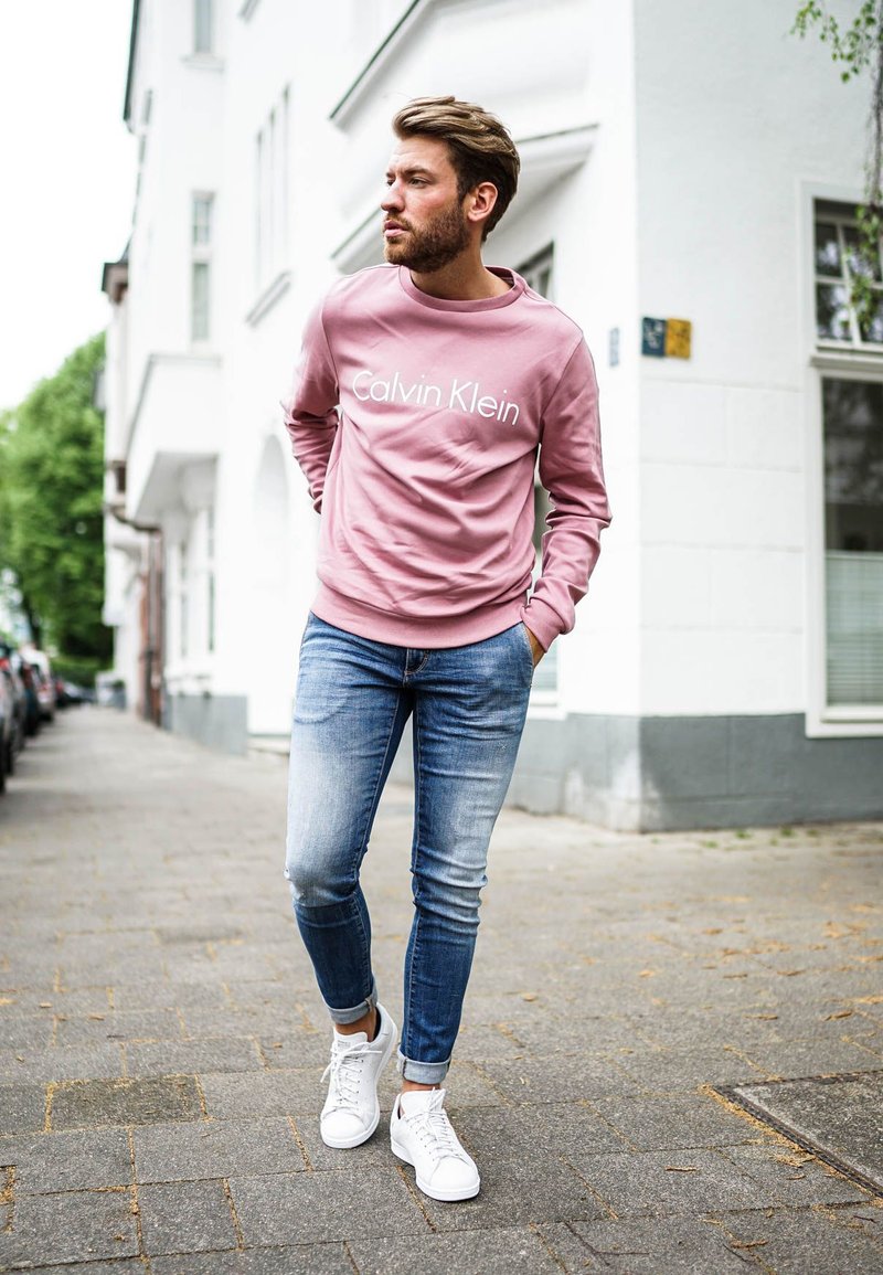 Rózsaszín kapucnis pulóver "Calvin Klein" logóval, kék farmer és fehér tornacipő, sétál a járdán fehér épületek mellett zöld növényzet között.