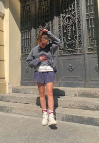 Sweatshirt cinza oversized, t-shirt branca por baixo, calções roxos, meias riscas vermelhas e sapatilhas brancas de cano alto. Escadas e porta decorativa ao fundo.