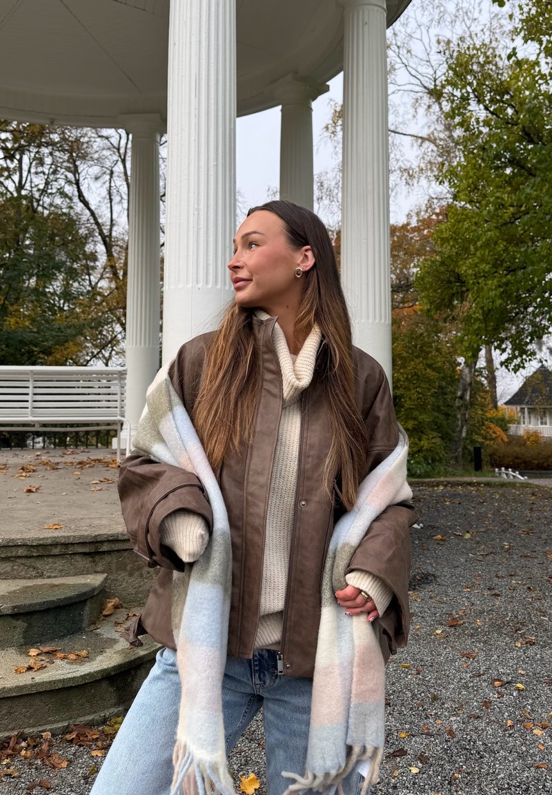 Brun læderjakke over en cremefarvet strikket sweater, dækket af et pastelternet tæppehalstørklæde. Blå jeans og en naturlig udendørs omgivelser.