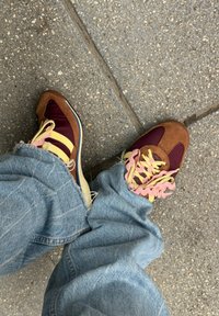 Bordo i smeđi velur tenisice sa žutim prugama i ružičastim vezicama, nosene s izlizanim plavim trapericama, postavljene na teksturiranom asfaltu.
