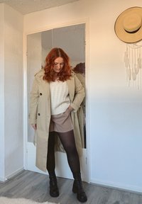 Beige trenchcoat, crèmekleurige gebreide trui, tan shorts, zwarte panty's en chunky heeled boots. Spiegels op de achtergrond, minimale decoratie.