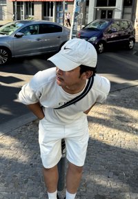 Λευκό πουκάμισο με μακριά μανίκια και ναυτικά διακοσμητικά, λευκές βερμούδες και λευκό καπέλο. Το άτομο στέκεται σε έναν λιθόστρωτο δρόμο κοντά σε παρκαρισμένα αυτοκίνητα.