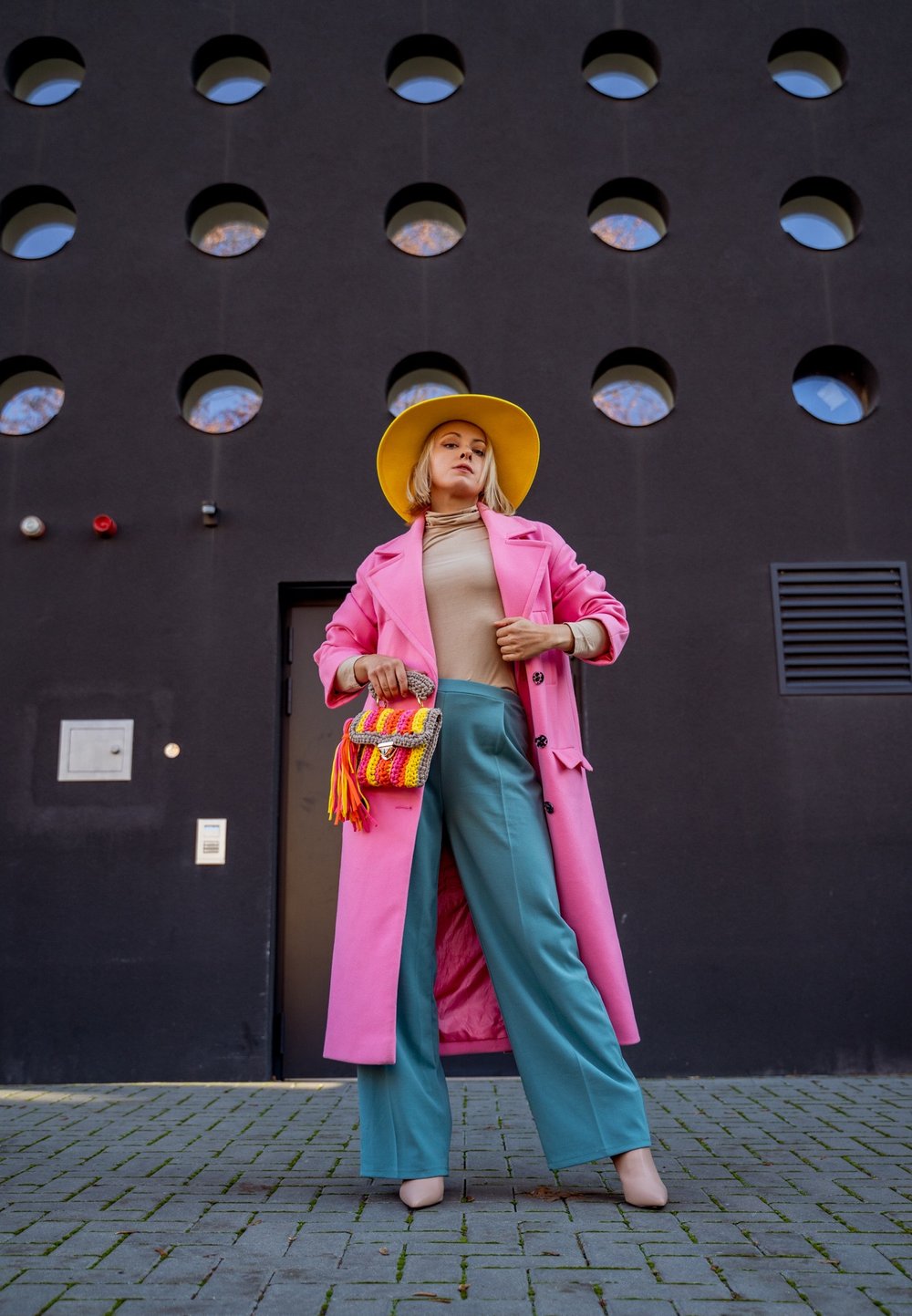 Ροζ αδιάβροχο, μπεζ ζιβάγκο, τιρκουάζ παντελόνι σε φάρδος, κίτρινο καπέλο, ζωντανή πολυχρωμία τσάντα με κρόσσια, σε αντίθεση με έναν σκοτεινό τοίχο με στρογγυλά παράθυρα.