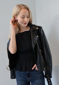 Zwarte leren motorjas met gouden accenten, gedragen over een zwarte textuurrijke top, gecombineerd met blauwe spijkerbroek. Tatoeages zichtbaar op de arm.