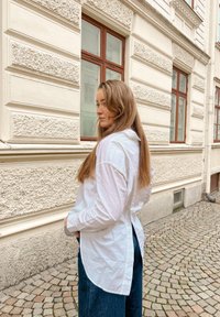 Biela dlhá mikina s kapucňou a rozparkom na chrbte, v kombinácii s tmavými rifľovými nohavicami. Na pozadí je textúrovaná svetlá stena.