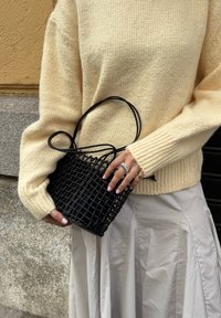 Bolsa de tecido preto com um design texturizado e alças duplas, segurada na mão, combinada com um suéter creme e uma saia de cor clara.