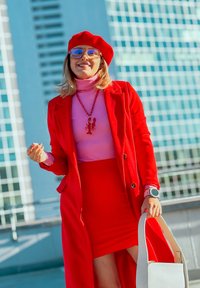 Červený kabát a ladící sukně, růžový rolák, červený baret. Brýle s průhlednými čočkami, náhrdelník s humrem. Bílá kabelka, růžové hodinky, uvolněná pozice.