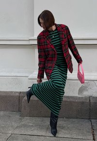 Red plaid blazer over a green striped maxi dress, paired with black heeled boots and a small pink handbag, set against a light background.