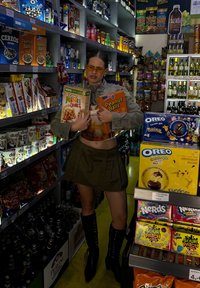 Dvije kutije žitarica drže se ispred polica punih grickalica i pića. Kutije su svijetlo narančaste i zelene boje, s veselim grafičkim dizajnom.