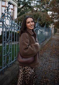 Young woman in brown fleece jacket and leopard print skirt stands smiling beside a wrought iron fence on a leaf-strewn sidewalk in autumn.