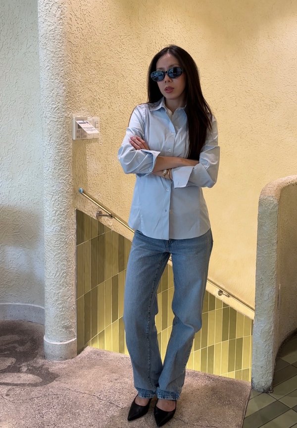Una mujer con gafas de sol, una camisa azul claro, vaqueros y zapatos negros de tacón está de pie con los brazos cruzados junto a un hueco de escalera alicatado.