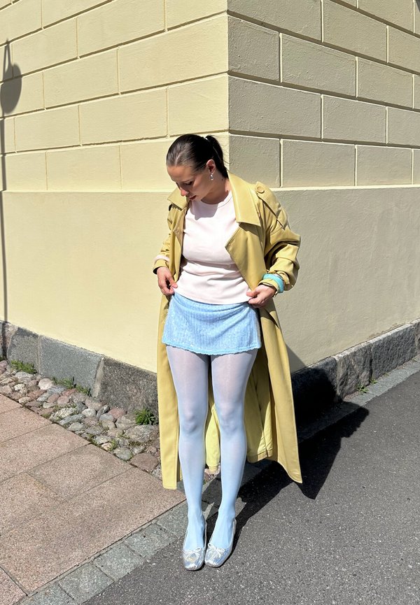 Vrouw die blauwe minirok rechtzet terwijl ze op een zonnige straathoek staat, gekleed in een gele jas, witte maillots en zilveren schoenen.