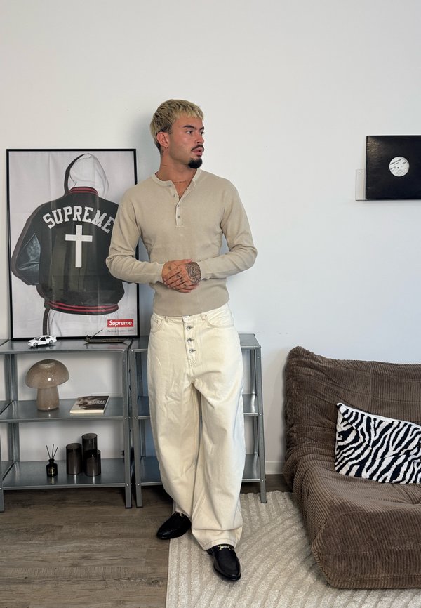 Mann med tatoveringer iført beige skjorte og hvite bukser står ved siden av en hylle med sopp-lampe, lekebil og innrammet bilde av en Supreme-jakke.