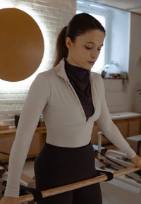 Nuori nainen treenivaatteissa pitää kädessään puista tankoa kirkkaassa, modernissa kuntosalissa, jossa taustalla on pilateslaitteita.