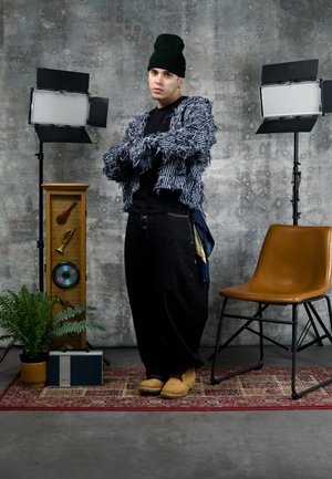 Joven con gorro negro, chaqueta de rayas y textura peluda, y pantalones anchos negros, de pie con los brazos cruzados entre luces de estudio, cerca de una silla, una planta y decoraciones musicales.