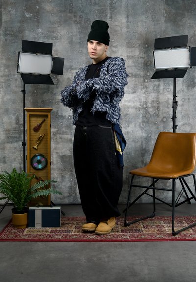 Joven con gorro negro, chaqueta de rayas y textura peluda, y pantalones anchos negros, de pie con los brazos cruzados entre luces de estudio, cerca de una silla, una planta y decoraciones musicales.