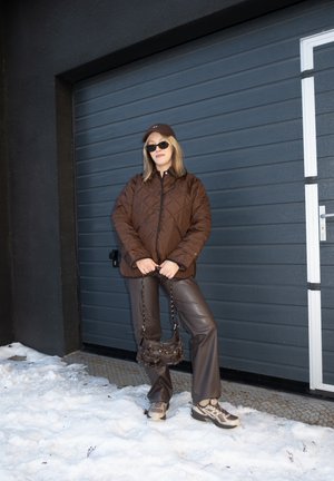 Mujer con chaqueta acolchada marrón, pantalones de cuero, zapatillas deportivas, gafas de sol y gorra, de pie sobre la nieve frente a una puerta de garaje oscura, sosteniendo una bolsa pequeña.