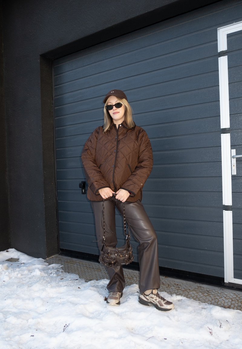Femme portant une veste matelassée marron, un pantalon en cuir, des baskets, des lunettes de soleil et une casquette, debout sur la neige devant une porte de garage sombre, tenant un petit sac.