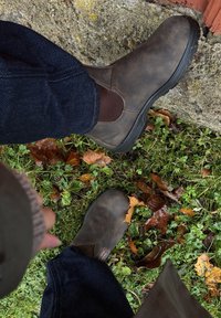 A person wearing dark brown boots and dark blue jeans stands on a mixture of grass, fallen leaves, and stone near a brick wall.