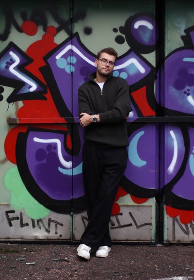 Suéter de punto verde oscuro de gran tamaño con una cremallera, pantalones negros y zapatillas blancas, frente a un fondo de graffiti colorido en tonos púrpura y rojo.