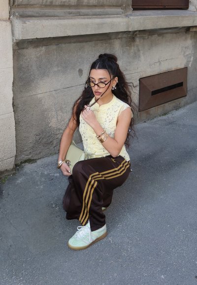 Mujer con el pelo largo, vestida con una camiseta sin mangas amarilla, pantalones marrones a rayas, zapatillas blancas y gafas, agachada en una acera junto a la pared de un edificio.