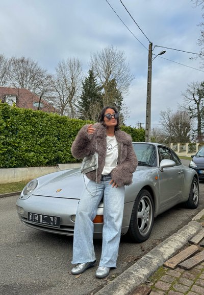 Mujer con chaqueta mullida y gafas de sol apoyada en un Porsche plateado estacionado en la calle, con árboles sin hojas y arbustos al fondo.