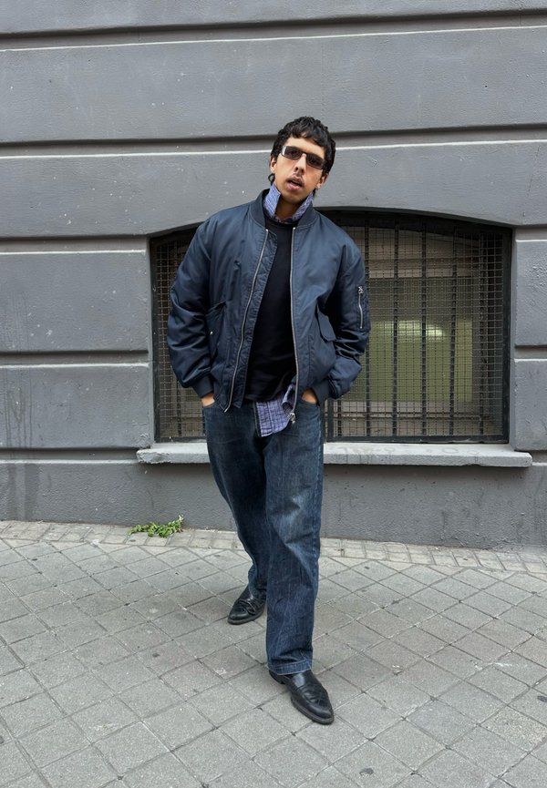 Homme portant des lunettes de soleil, une veste bomber bleu marine, une chemise bleue et un jean, debout les mains dans les poches contre un mur gris avec une fenêtre grillagée.