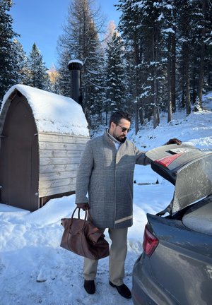 Hombre con abrigo y gafas de sol sosteniendo una bolsa de lona, cargando objetos en el maletero de un coche en un bosque nevado cerca de una pequeña cabaña de madera.