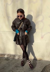 Manteau en cuir marron avec une ceinture, collants noirs et chaussures bordeaux. Comprend des poignets bleus et un serre-tête à pois. Debout contre un mur.