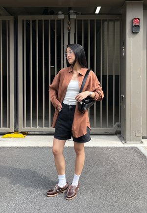 Camisa de lino marrón de botones, tank top blanco, pantalones cortos negros, calcetines blancos, zapatos marrones, de pie cerca de puertas de metal. Materiales texturizados mencionados.