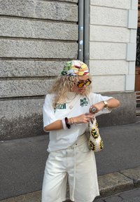 Vitt t-shirt med grön text, krämfärgade shorts, mönstrad huvudduk, gula solglasögon och en handgjord väska med gröna och gula detaljer.