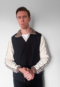 Veste de sport noire et crème avec un col marron, fermeture par boutons et accents rayés sur les manches. Texture lisse, coupe ajustée.