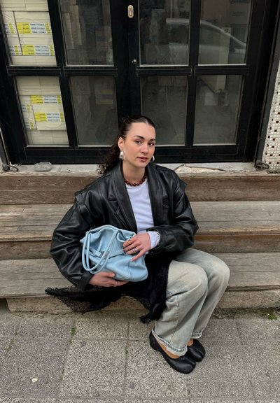 Mujer con el cabello oscuro recogido, vestida con chaqueta de cuero negra y jeans claros, sentada en escaleras de madera sosteniendo un bolso azul claro.
