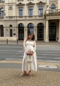 Mlada žena v beli čipkasti obleki drži torbico z biseri in sončna očala, stoji na pločniku blizu prehoda za peščce ob zgodovinski stavbi.