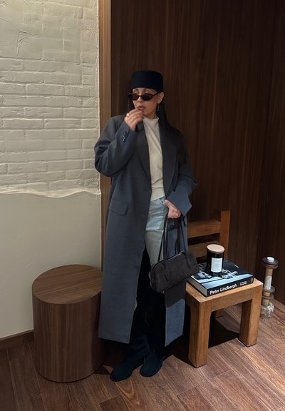 Mujer con abrigo largo gris, suéter claro, pantalones claros, sombrero negro y gafas de sol, sosteniendo un bolso negro y recostada contra una pared de madera junto a un taburete y una mesa pequeña.