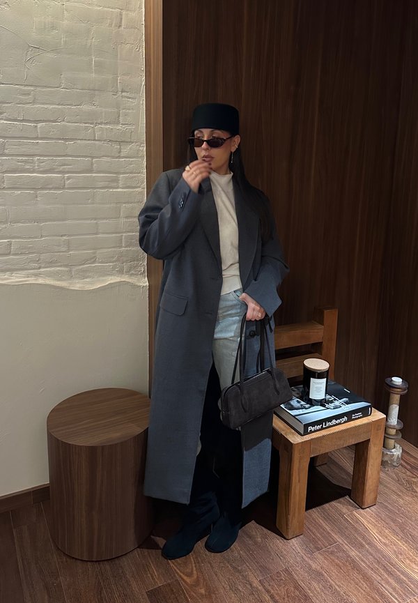 Kvinna i lång grå kappa, ljus tröja, ljusa byxor, svart hatt och solglasögon som håller en svart väska och lutar sig mot en trävägg nära en pall och ett litet bord.