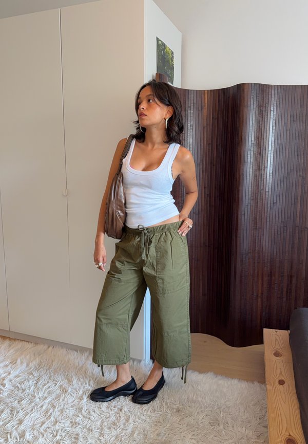 Mujer con camiseta blanca sin mangas, pantalones anchos color verde oliva, zapatos negros y llevando un bolso marrón al hombro, de pie sobre una alfombra blanca y peluda en interiores.