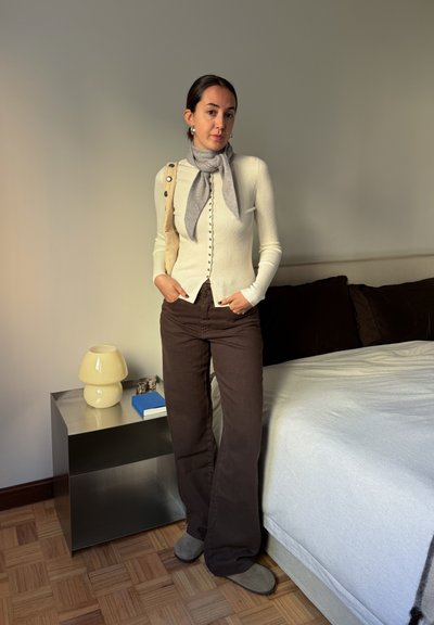 Cárdigan de punto color crema con detalles de botones, bufanda gris, pantalones acampanados marrones y calzado gris. Decoración minimalista con una lámpara y una mesita de noche.