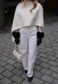 Personne portant un pantalon blanc à jambes larges, une cape crème, des gants noirs avec des poignets en fourrure, des chaussures noires pointues, et tenant un petit sac à main crème.