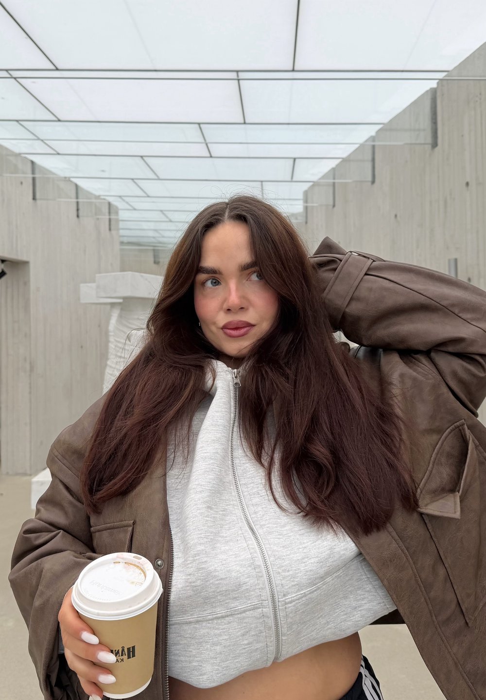 Brązowa skórzana, oversize'owa kurtka na szarej bluzie z kapturem zapinanej na zamek, trzymająca kubek kawy z beżowym rękawem, na tle minimalistycznej scenerii na zewnątrz.