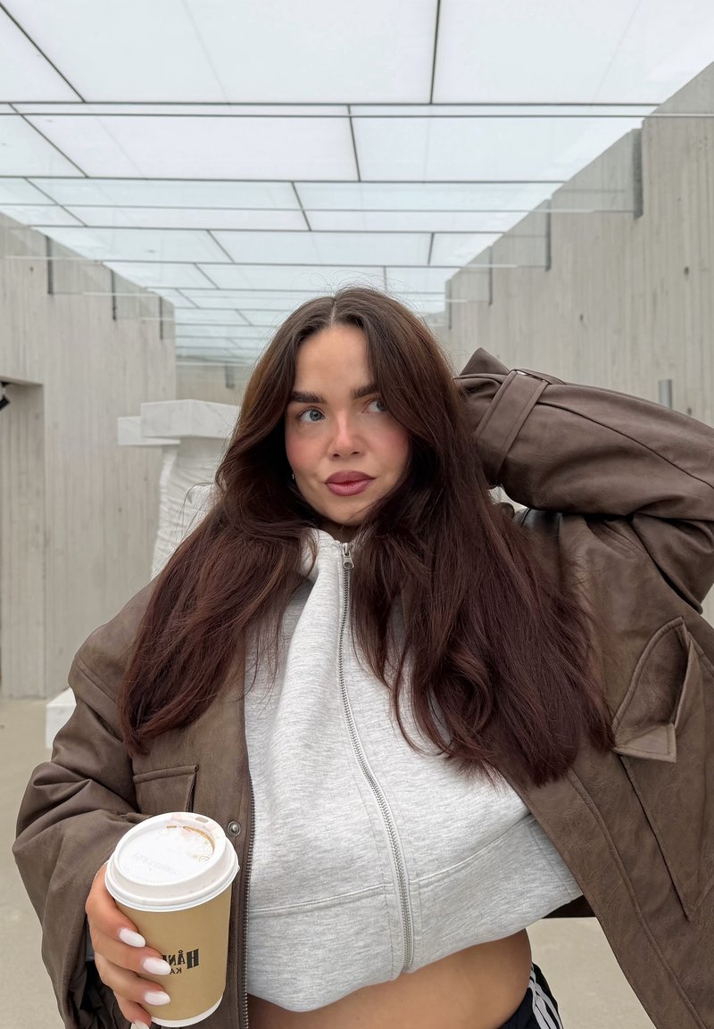 Brown leather oversized jacket over a gray zip-up hoodie, holding a coffee cup with a beige sleeve, against a minimalist outdoor setting.