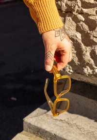 Des lunettes de soleil surdimensionnées jaunes avec des verres foncés sont tenues dans une main. La monture est transparente, avec un design géométrique carré.