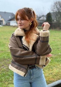 Femme rousse en extérieur portant une veste en peau retournée marron, un jean bleu, des boucles d'oreilles dorées, les cheveux attachés en queue de cheval, regardant vers la gauche.